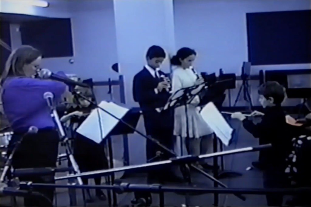 Blue-tinted vintage video still of children performing on recorders and other instruments at music stands.