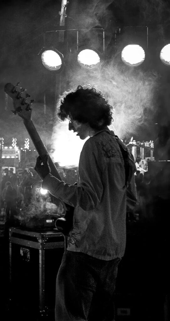 Black-and-white live shot: silhouette of Zack Rosen playing bass guitar on stage, backlit by stage lights and smoke.