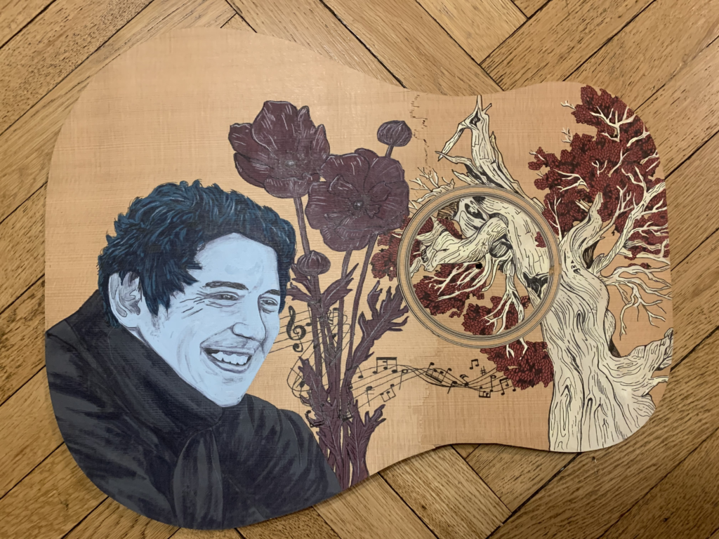 A guitar soundboard on a herringbone wood floor, hand-painted with a portrait of a smiling curly-haired young man beside dark poppies, musical notes, and a gnarled tree.