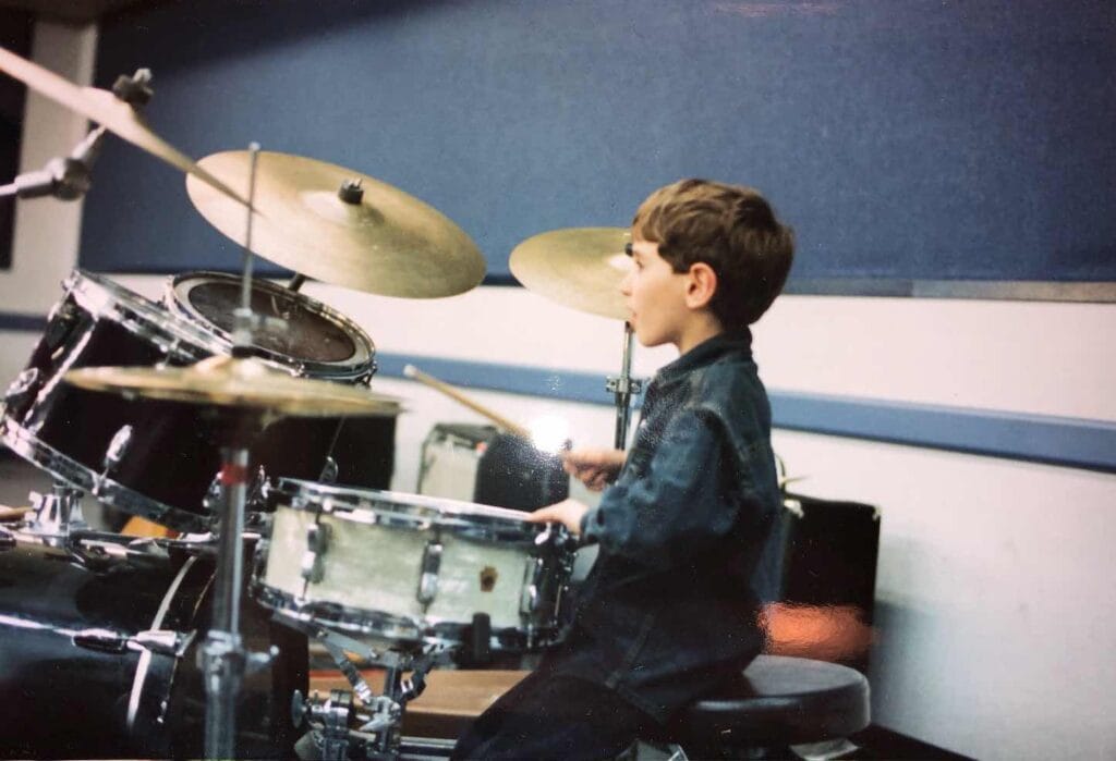 Vintage photo of Zack Rosen as a young boy in a denim shirt, mid-strike on a drum kit.