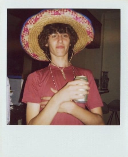 Polaroid of Zack Rosen in a straw sombrero and beaded necklace, arms crossed, holding a drink can indoors.
