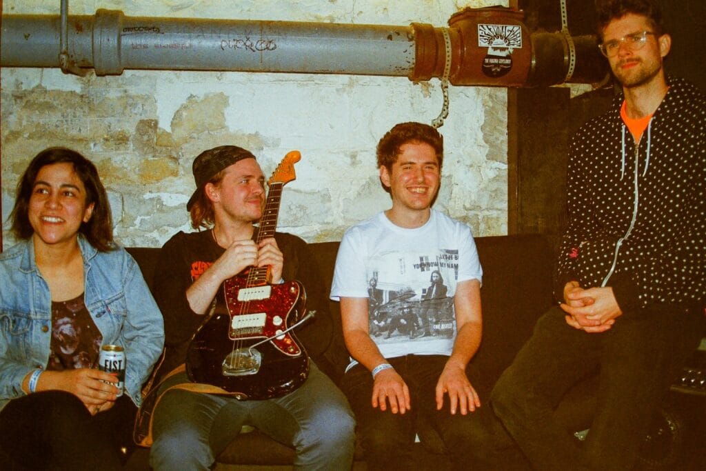 Four young people on a couch against an exposed-brick and pipe wall; one holds an electric guitar, one a drink can.