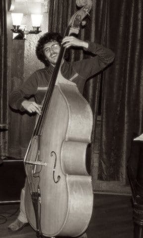 Sepia photo of Zack Rosen, smiling, with glasses and curly hair, playing an upright double bass in a dim room.
