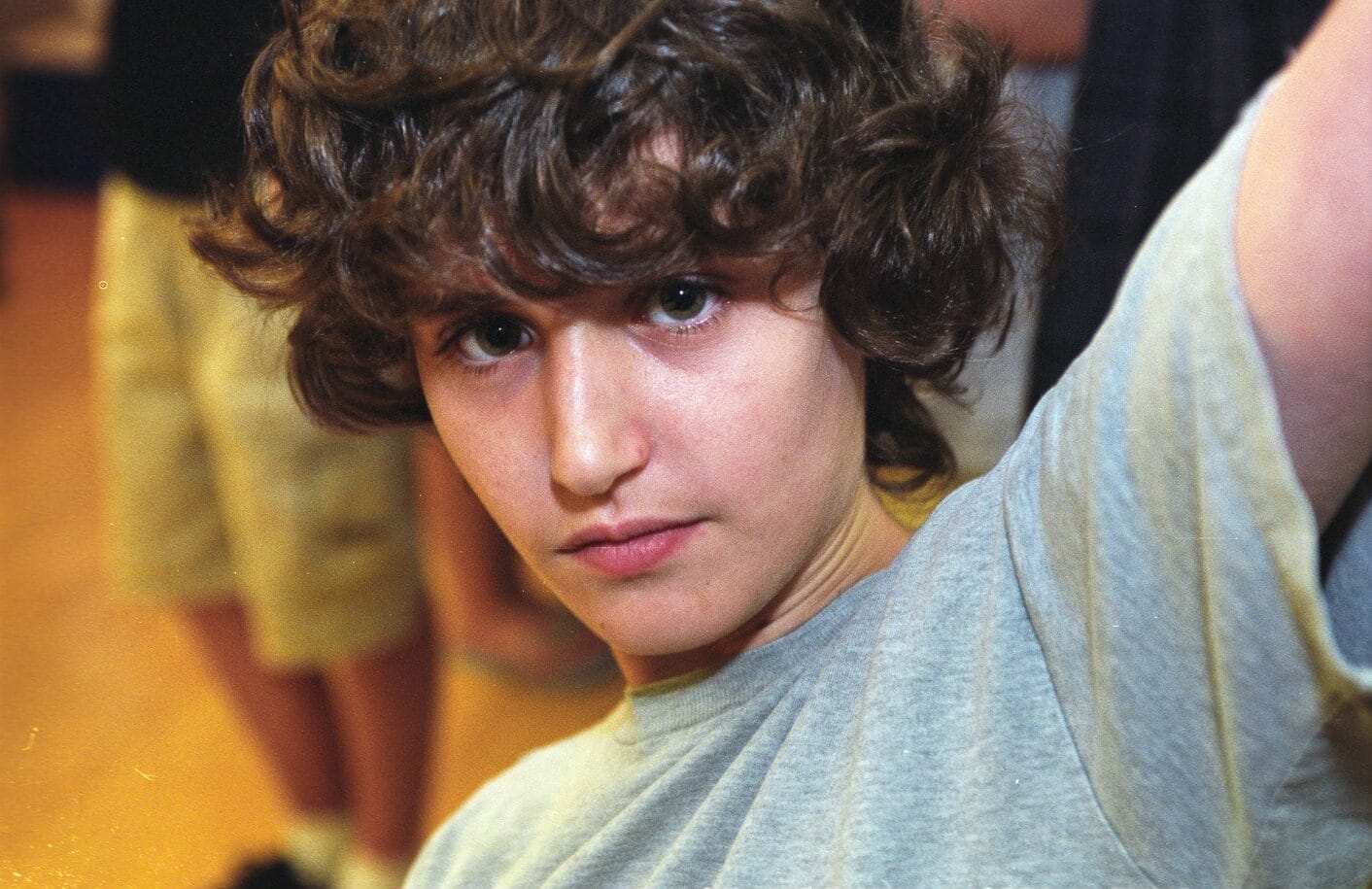 Close-up color portrait of Zack Rosen — curly brown hair, grey T-shirt — arm raised, looking toward the camera; warm indoor background.