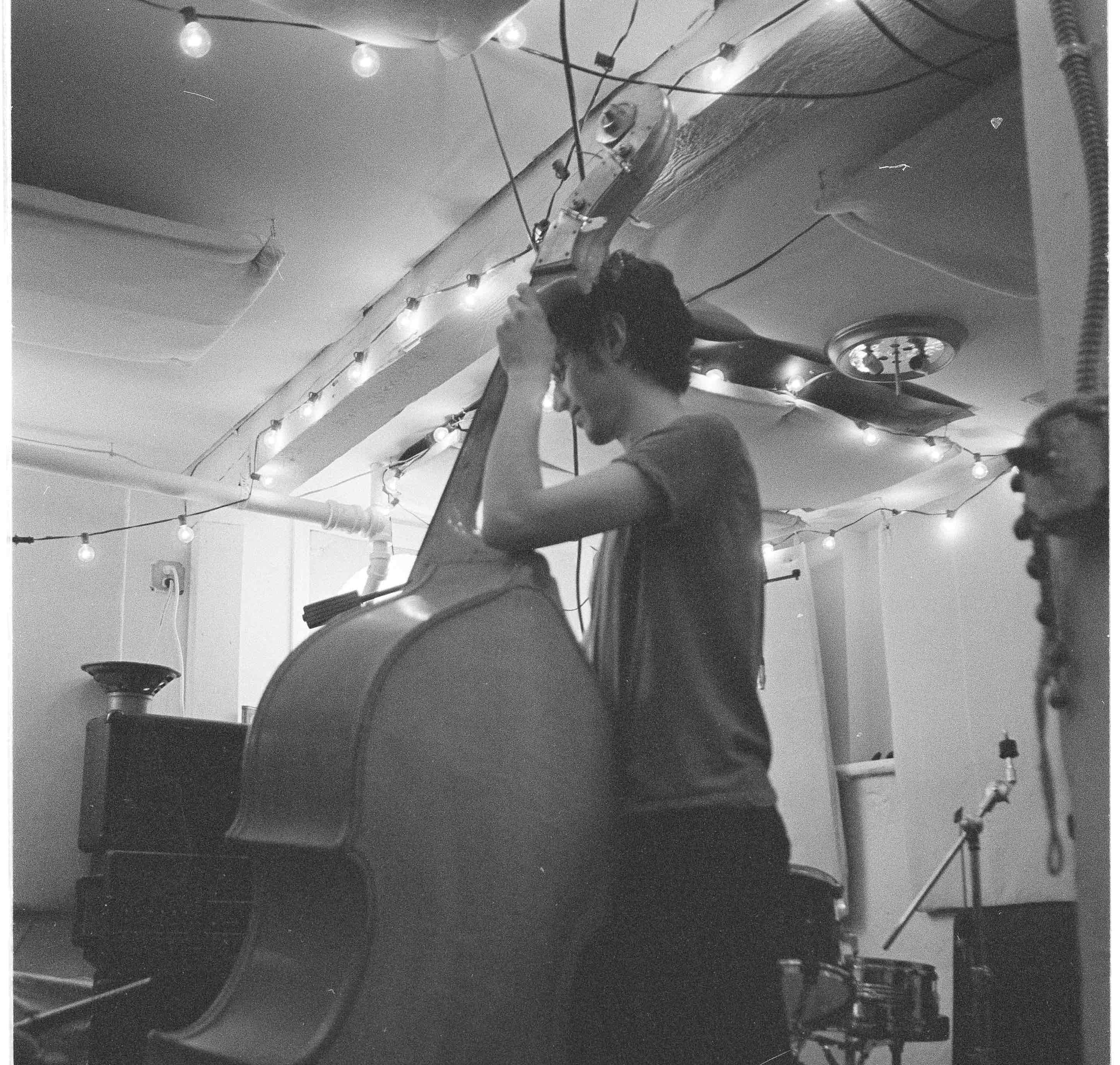 Black-and-white photo of Zack Rosen playing an upright double bass in a room strung with string lights.