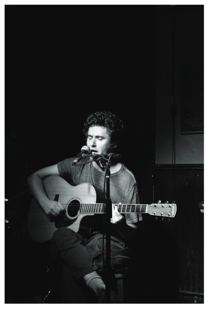 Black-and-white photo of Zack Rosen on a stool, singing into a microphone while playing acoustic guitar in a dim venue.