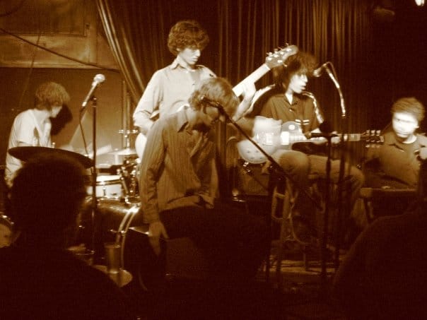 Sepia photo of the band Flearoy performing — singer at a mic, drummer, and guitarist — before a seated audience.